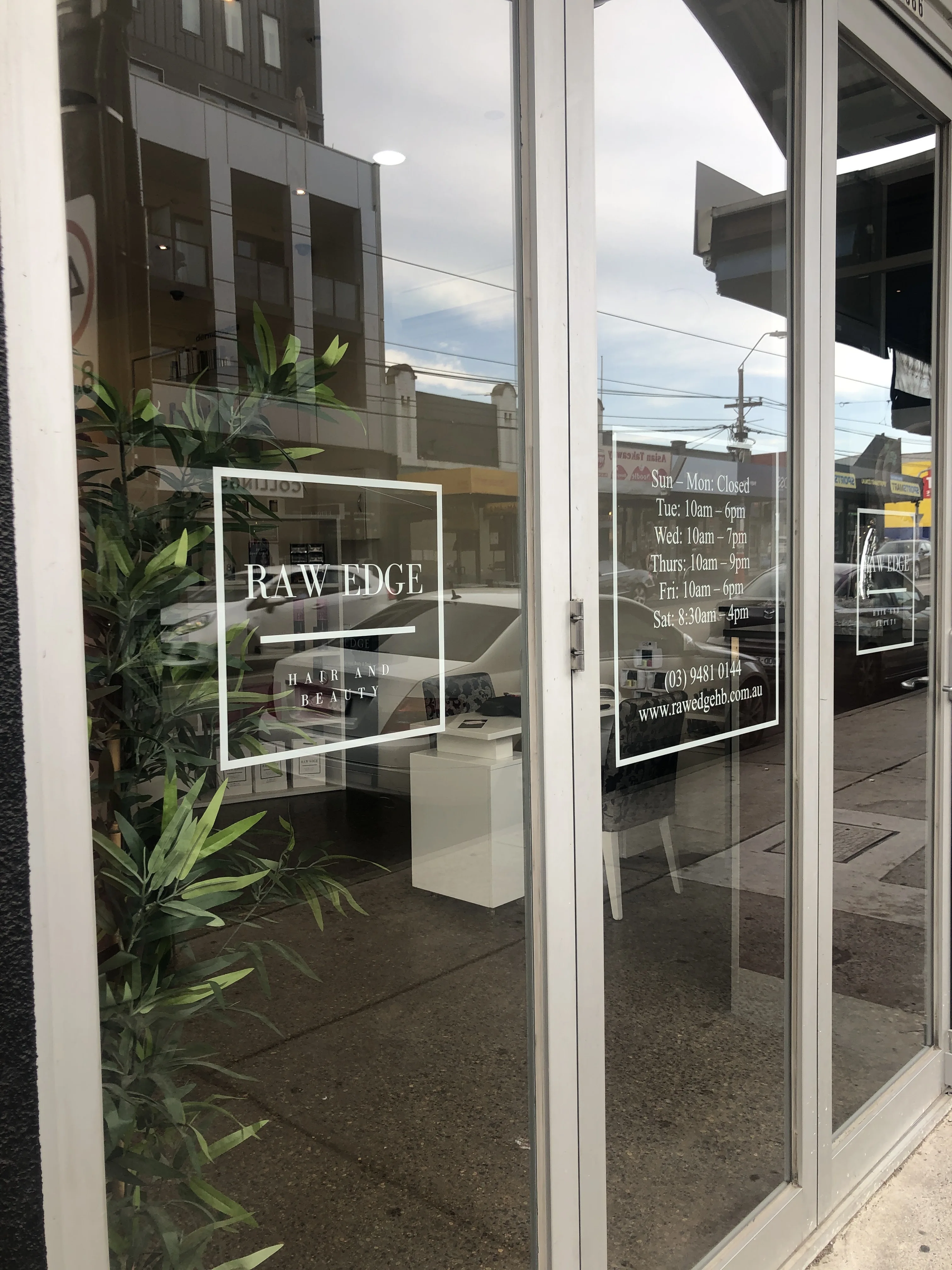 Raw Edge shopfront glass in portrait — wordmark, trading hours and phone number etched in the window