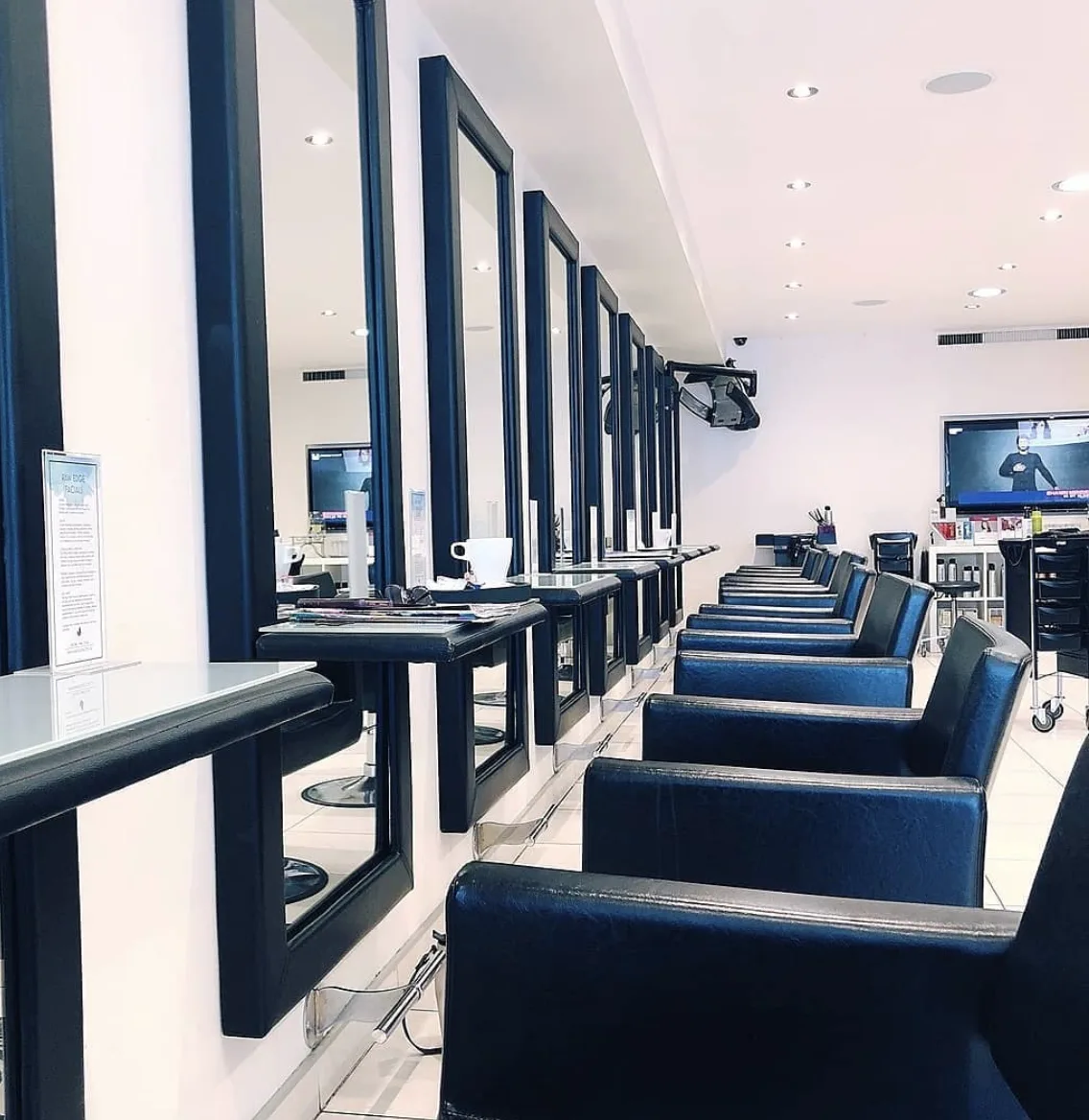 Raw Edge salon interior — receding row of black-framed mirrors and black leather styling chairs, white-tiled floor